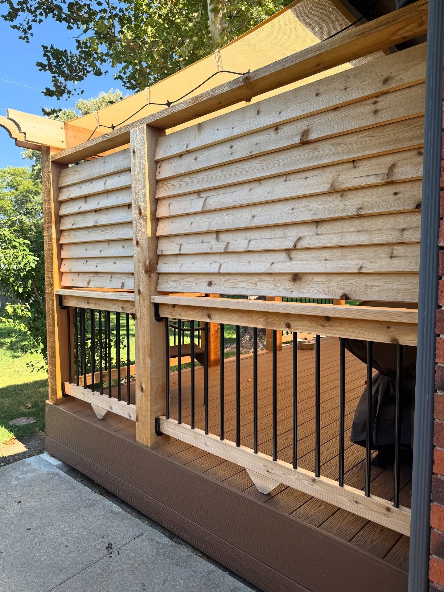 A wooden deck partition with horizontal slats above black metal railing, set against a brick wall and outdoor green space.