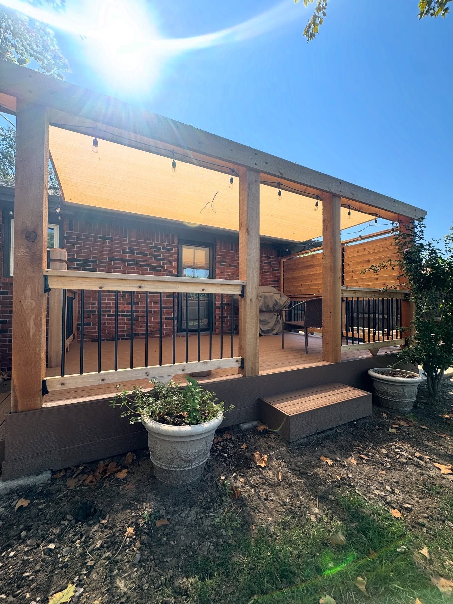 A sunlit wooden deck featuring a tan shade sail, black railings, and potted plants against a red brick house exterior.
