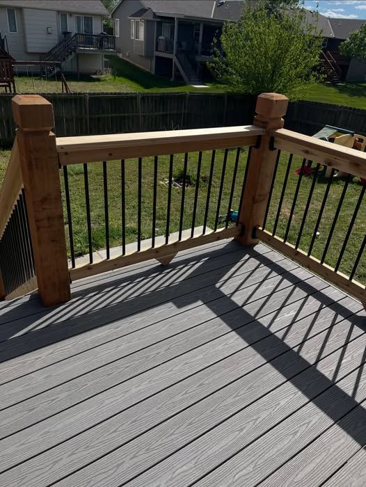 A grey composite deck with wooden posts and black metal balusters overlooking a fenced backyard on a sunny day.