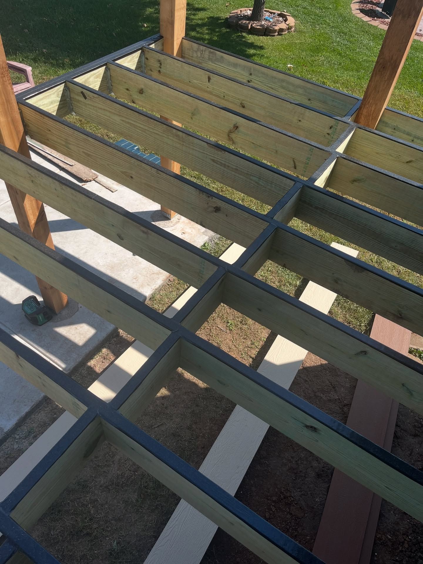Wooden deck frame under construction with joists lined with black protective tape, set on a grass and concrete backyard.