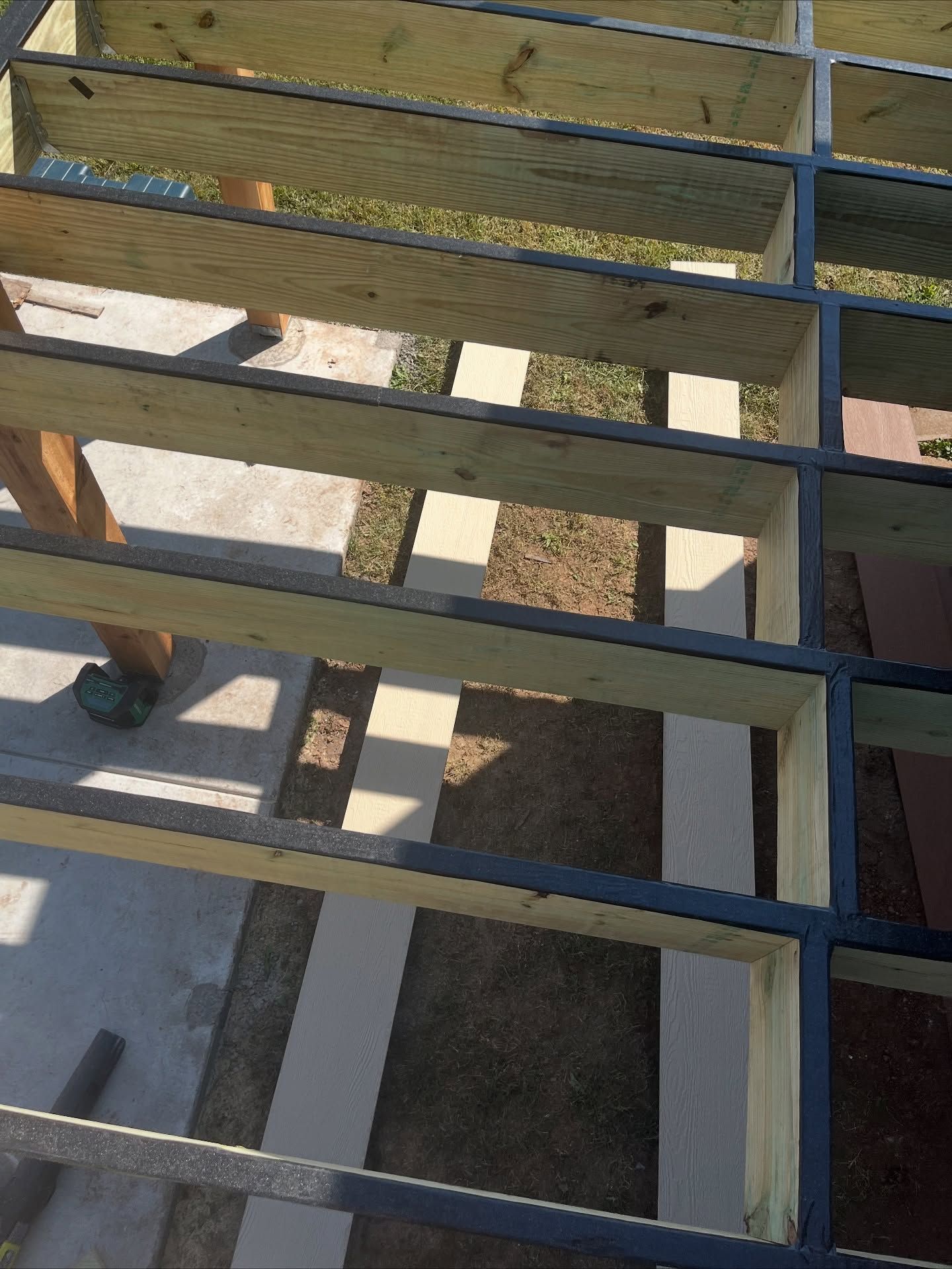 A high-angle view of wooden deck framing with dark protective joist tape applied to the top edges of the lumber.
