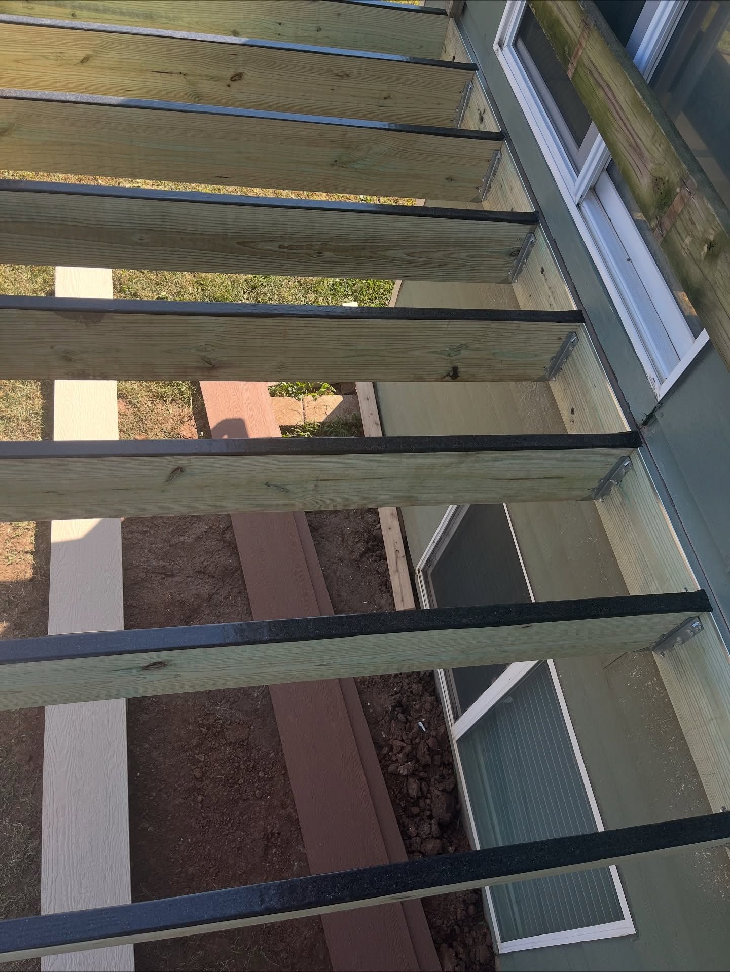A top-down view of deck joists with black protective tape applied to the top edges, installed alongside a house wall.