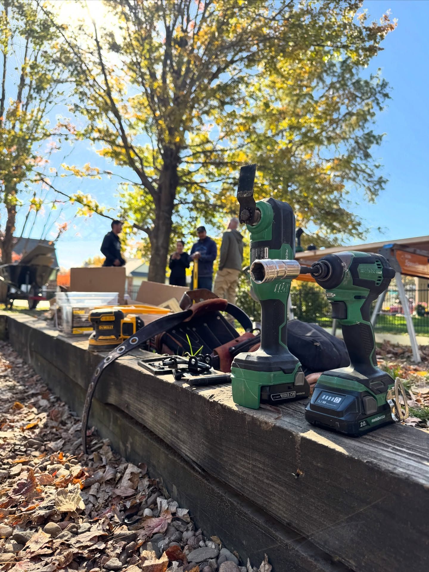 Green cordless power tools sit on a wooden beam outdoors, with several people standing in the background under trees.