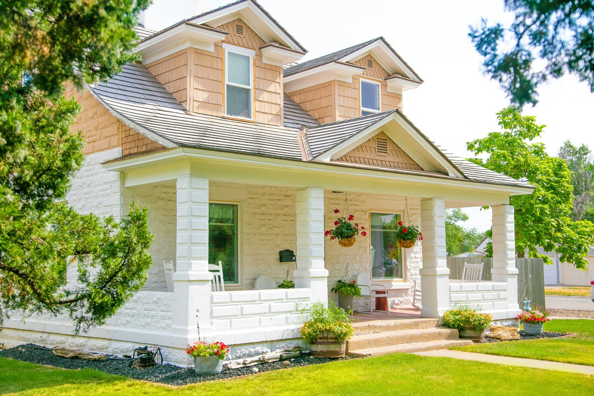 Two-story white brick house with a covered porch and dormers, set on a green lawn with trees.