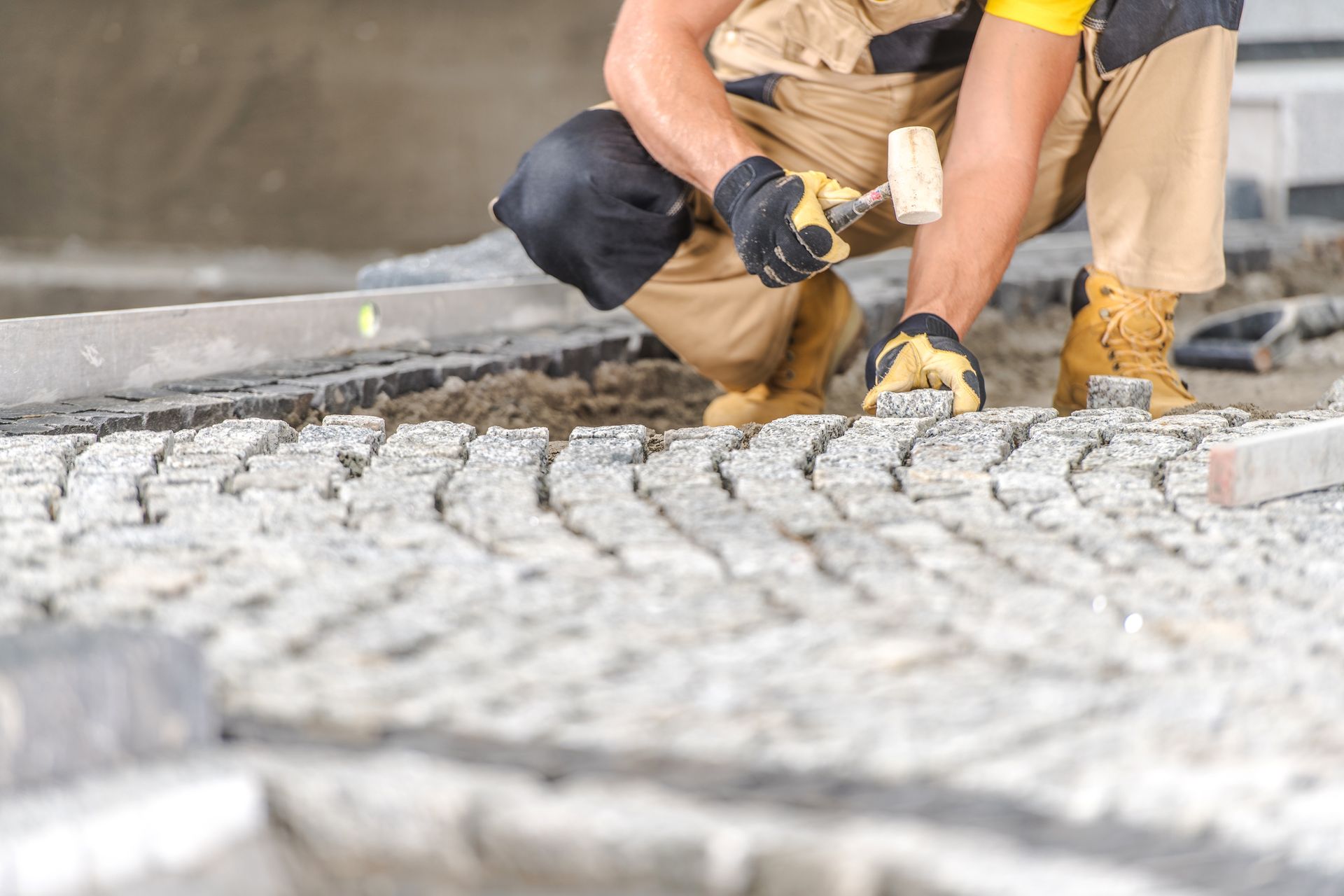A man is kneeling down while using a hammer to hammer bricks on a sidewalk.