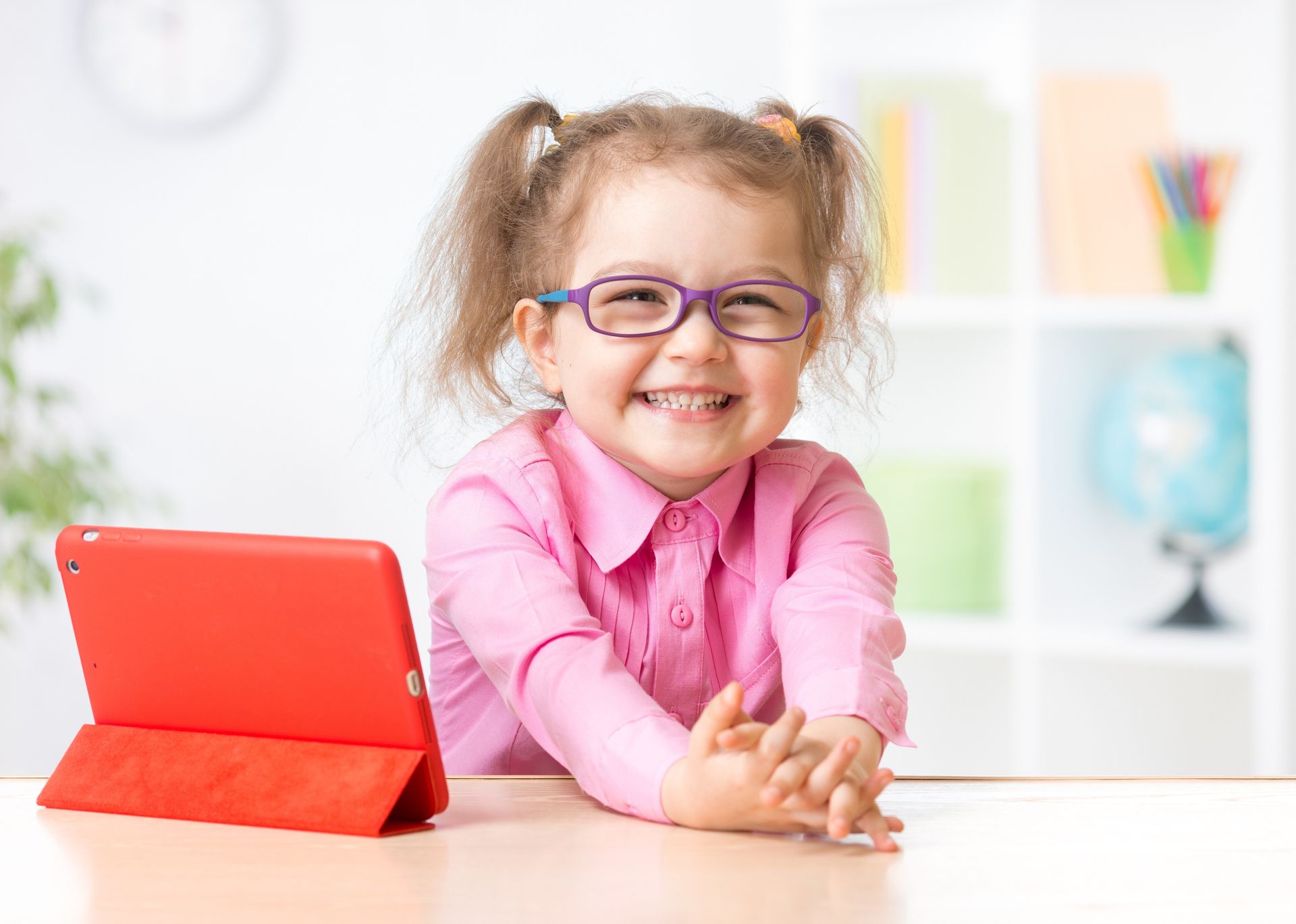 A little girl wearing glasses is sitting at a table with a tablet.