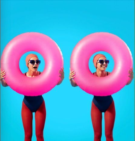 Two women in swimsuits are wearing woow sunglasses and 
holding pink inner tubes over their heads.