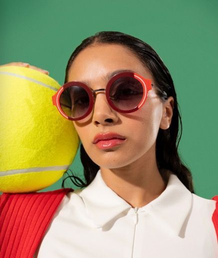 A woman wearing  woow sunglasses is holding a tennis ball on her shoulder.