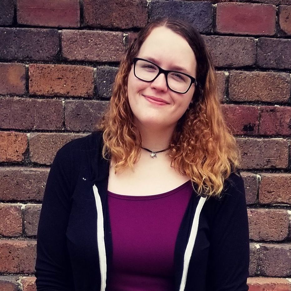 Woman with glasses smiles in front of a brick wall, wearing a black jacket over a purple shirt.
