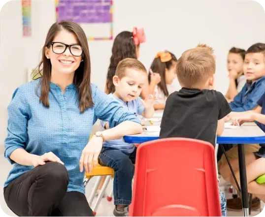 A teacher is sitting in front of a group of children sitting at tables in a classroom.