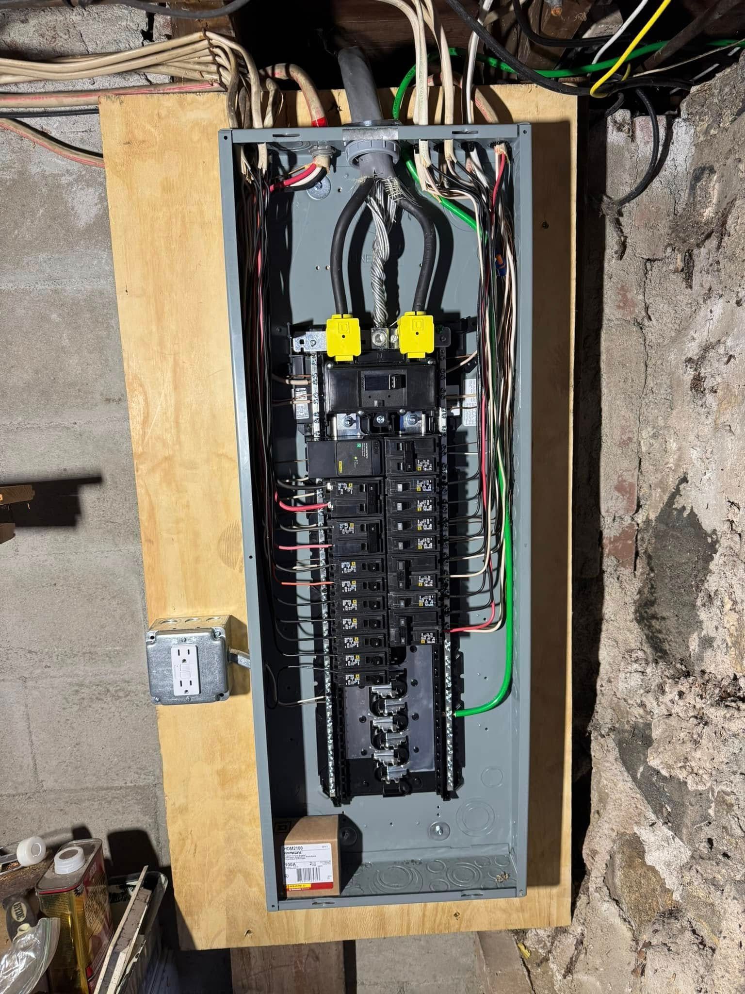 Electrical panel mounted on wood in a basement. Wires feed into panel with breakers; a wall outlet is visible to the left.
