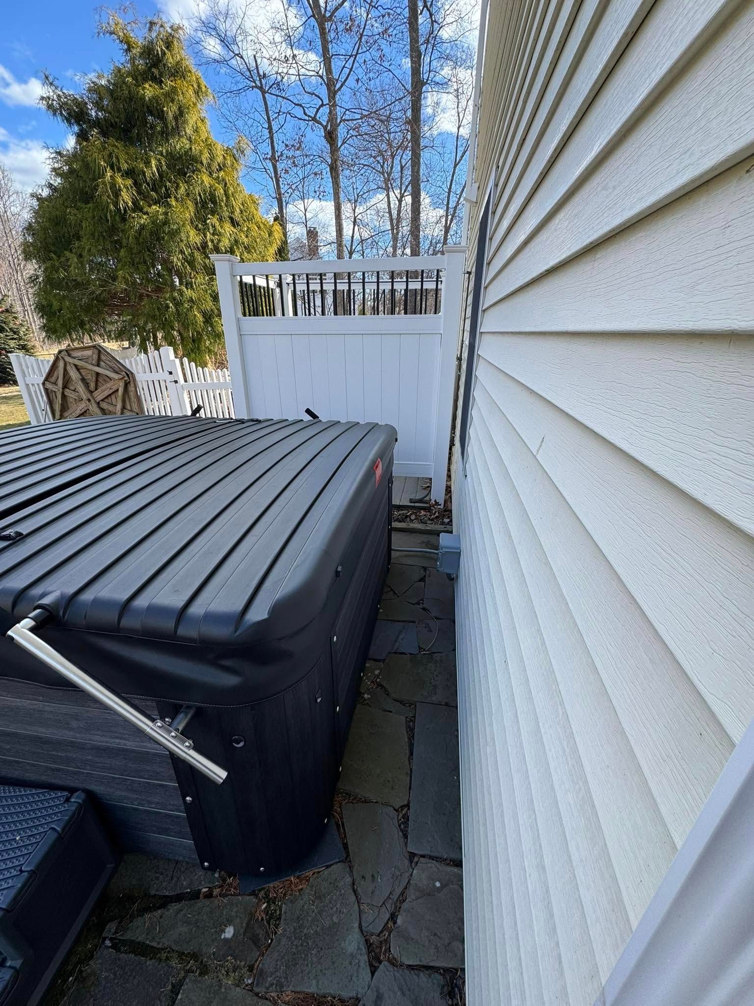 Black hot tub next to a white house with a white fence, set on a stone patio.