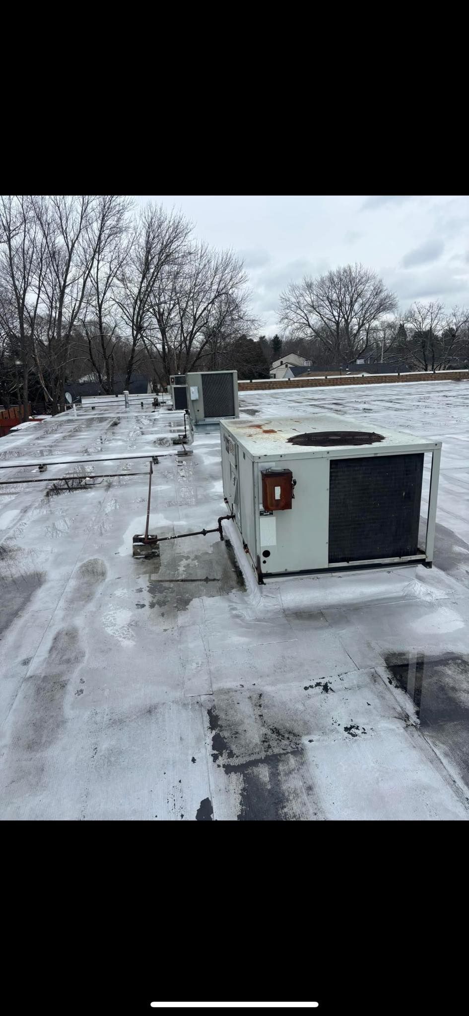 A snowy rooftop with HVAC units. Trees and overcast sky in the background.