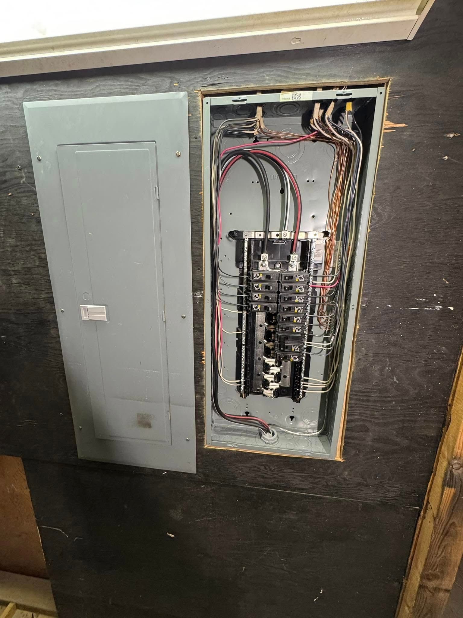 Electrical panel with door open, revealing wiring and circuit breakers, mounted on a dark wall.