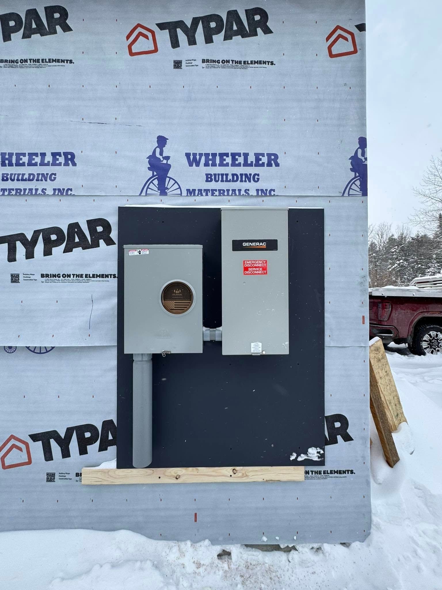Exterior electrical panel and meter box on a building under construction, mounted on a dark backer board; snow surrounds.