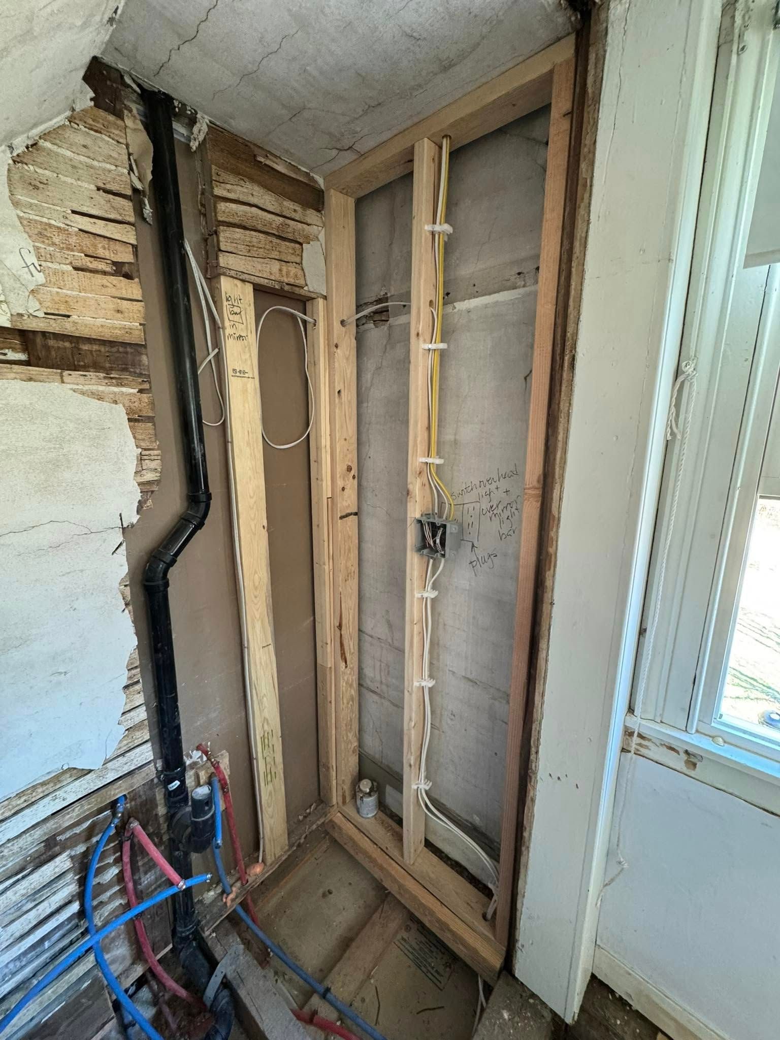 Bathroom under construction: exposed framing, pipes, and electrical wiring. A window is on the right.