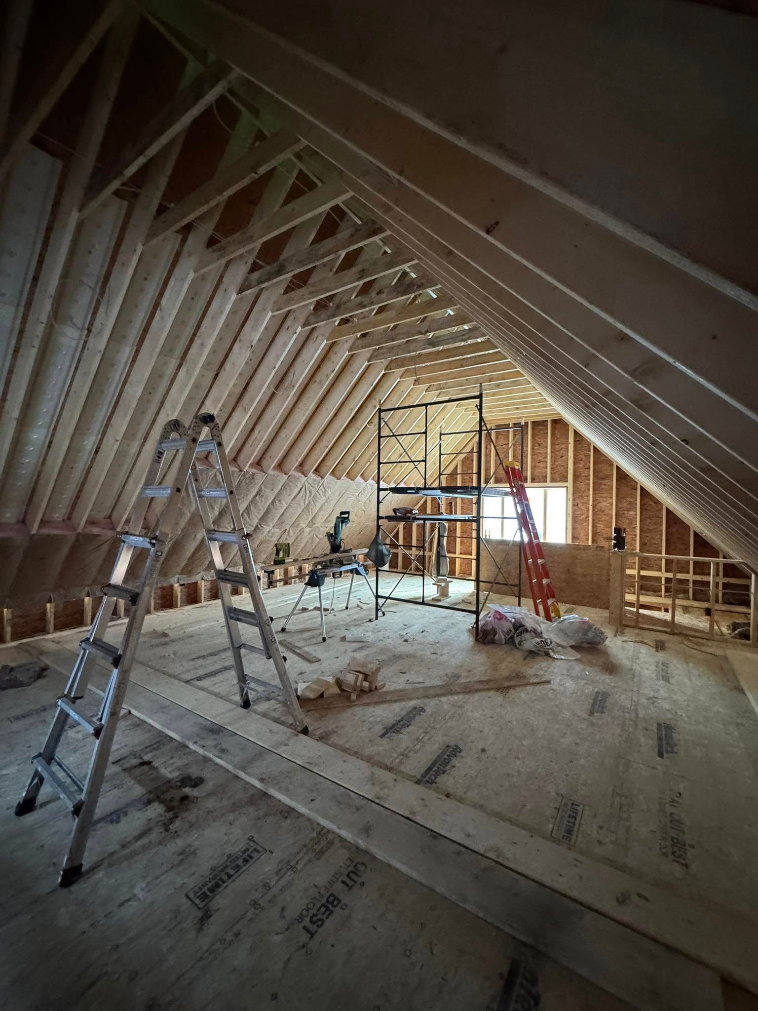 Interior of a building's attic under construction with wooden framing, ladders, and exposed studs.