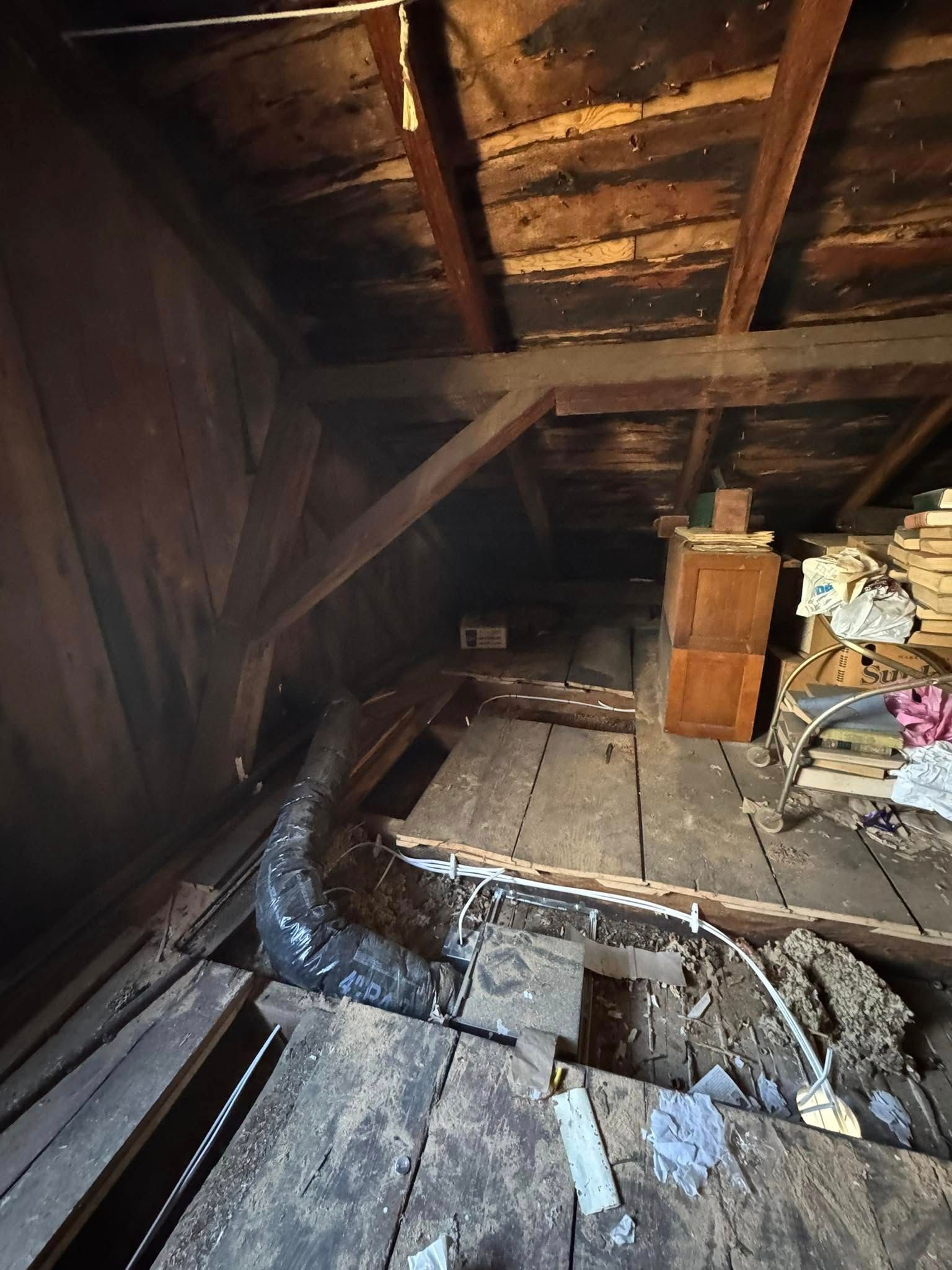 Interior attic view: wooden beams, a chimney, and a dark metal pipe against a dim, cluttered space.