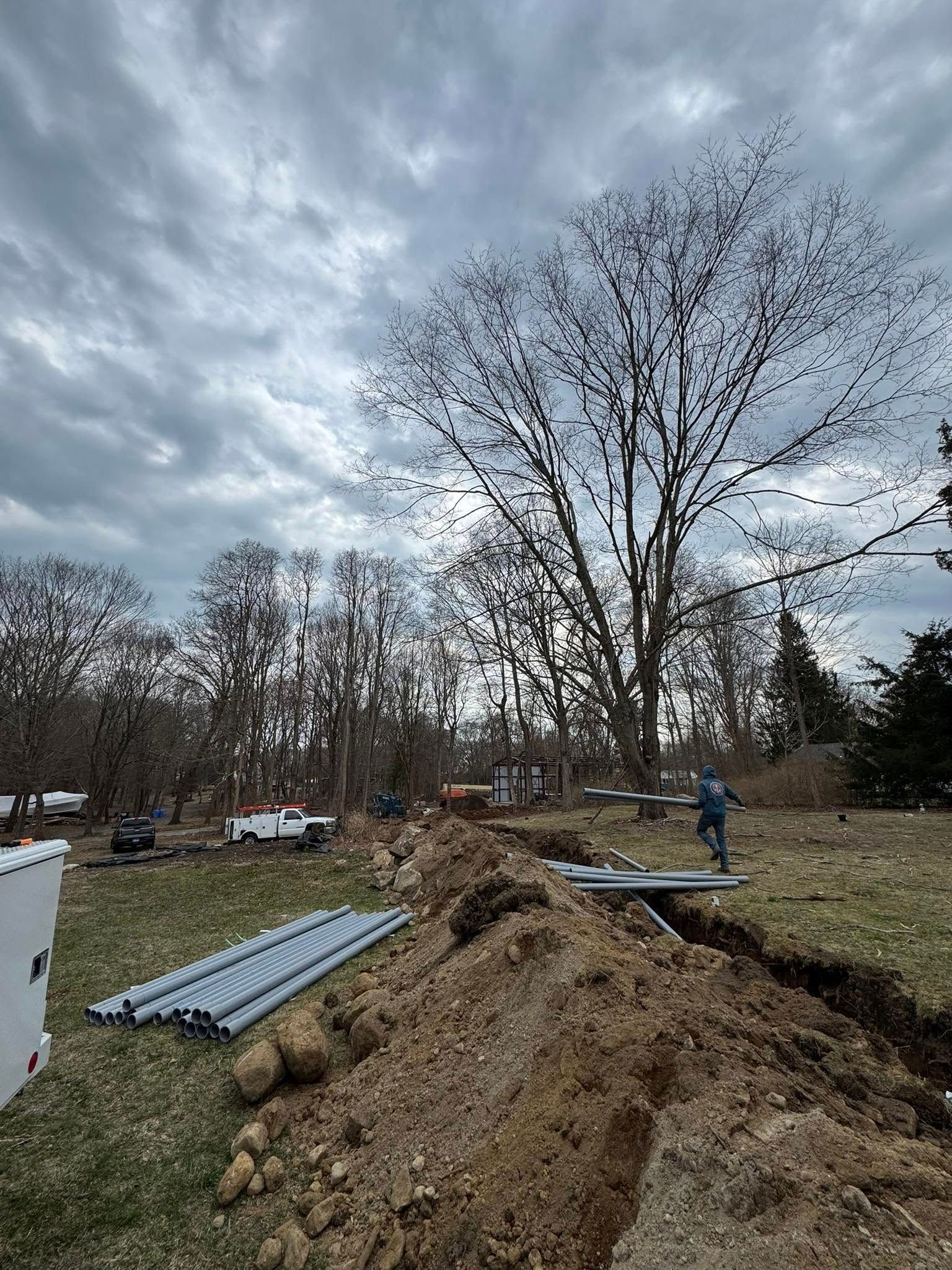 Construction site with a man, dirt mound, pipes, and trees under a cloudy sky.