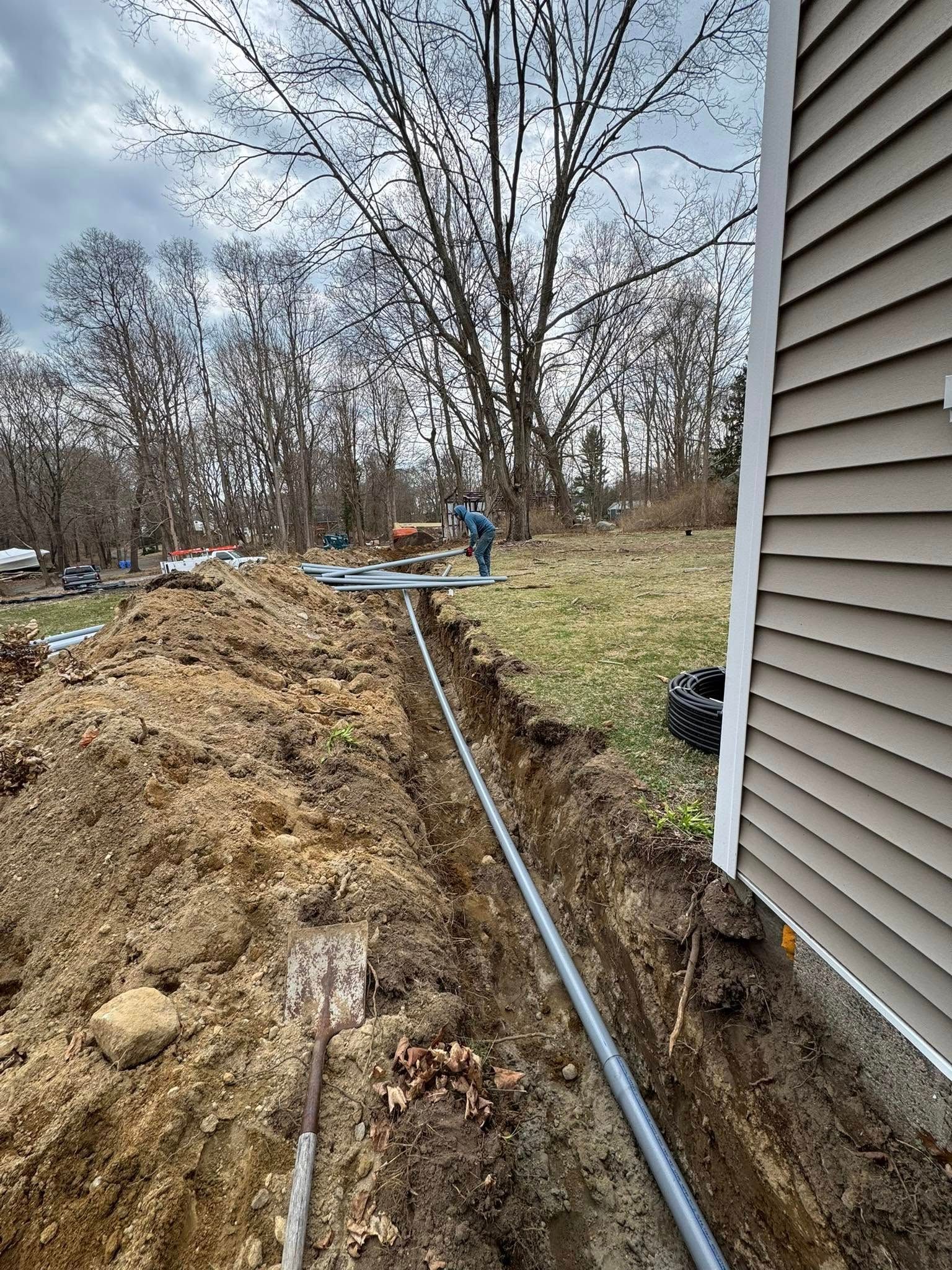 Trench dug alongside a house, with conduit pipe laid. Shovel sits in the foreground, worker in the distance.
