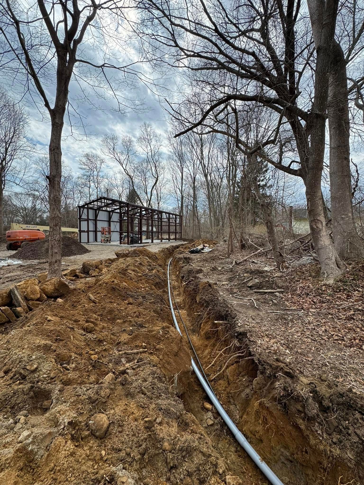 A trench with gray pipe leads to a building frame construction site. Trees surround the area on a cloudy day.