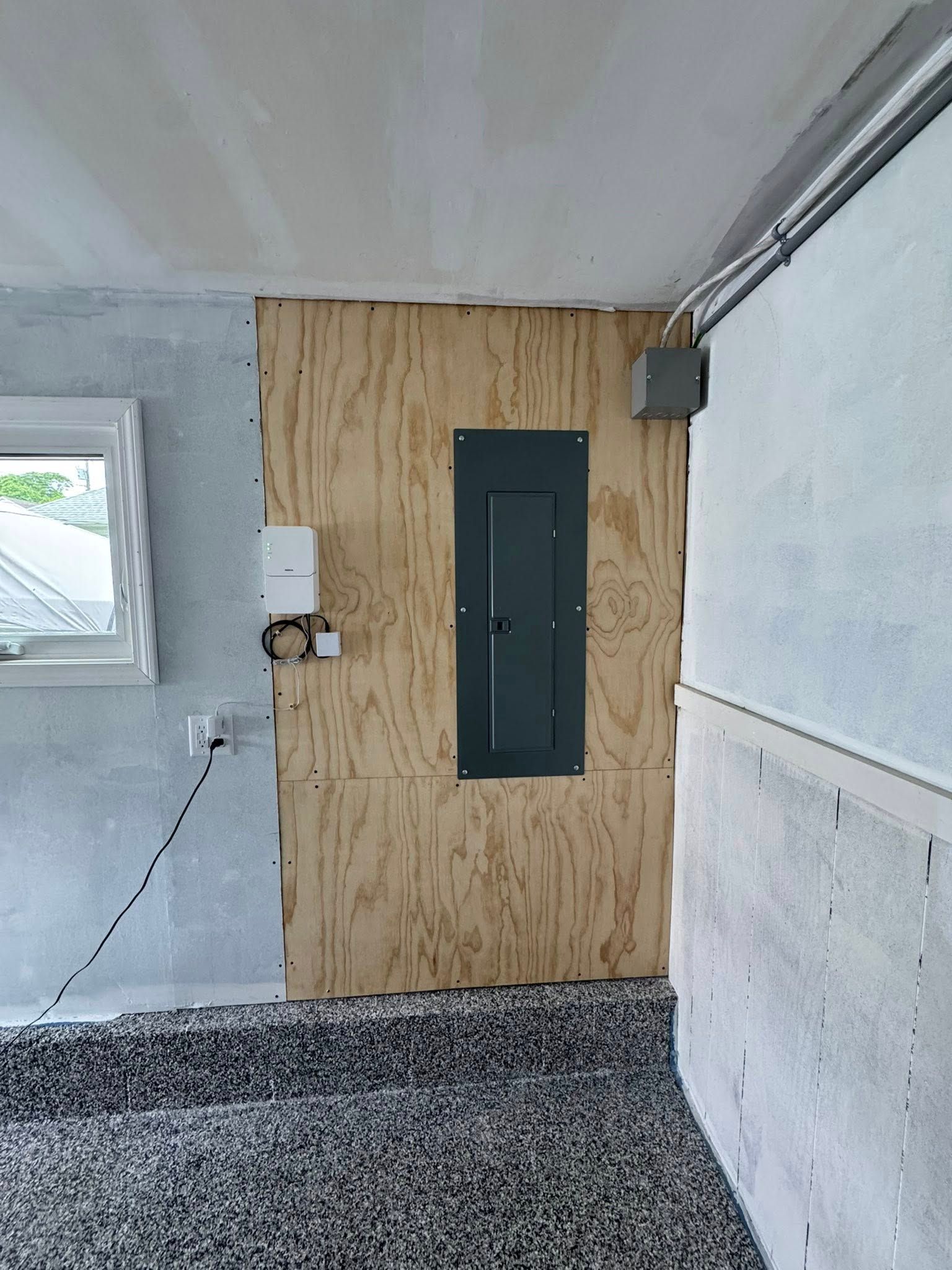 Electrical panel mounted on plywood in a room with unfinished walls and flooring.