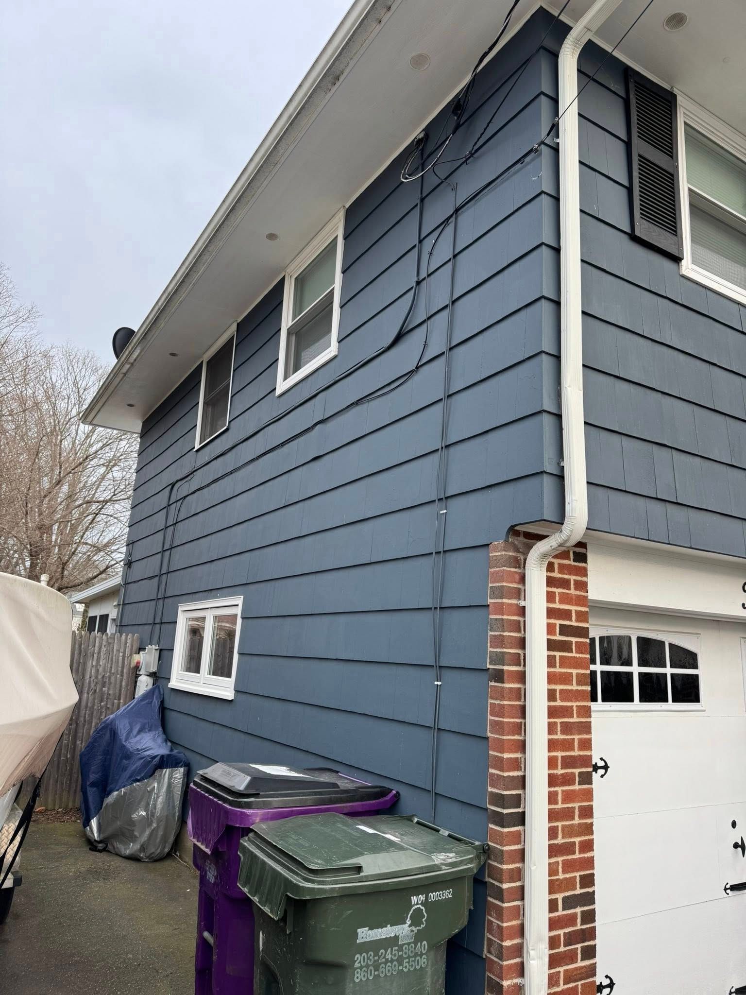 Side view of a two-story blue-sided house with a white garage door, trash bins, and utility wires.
