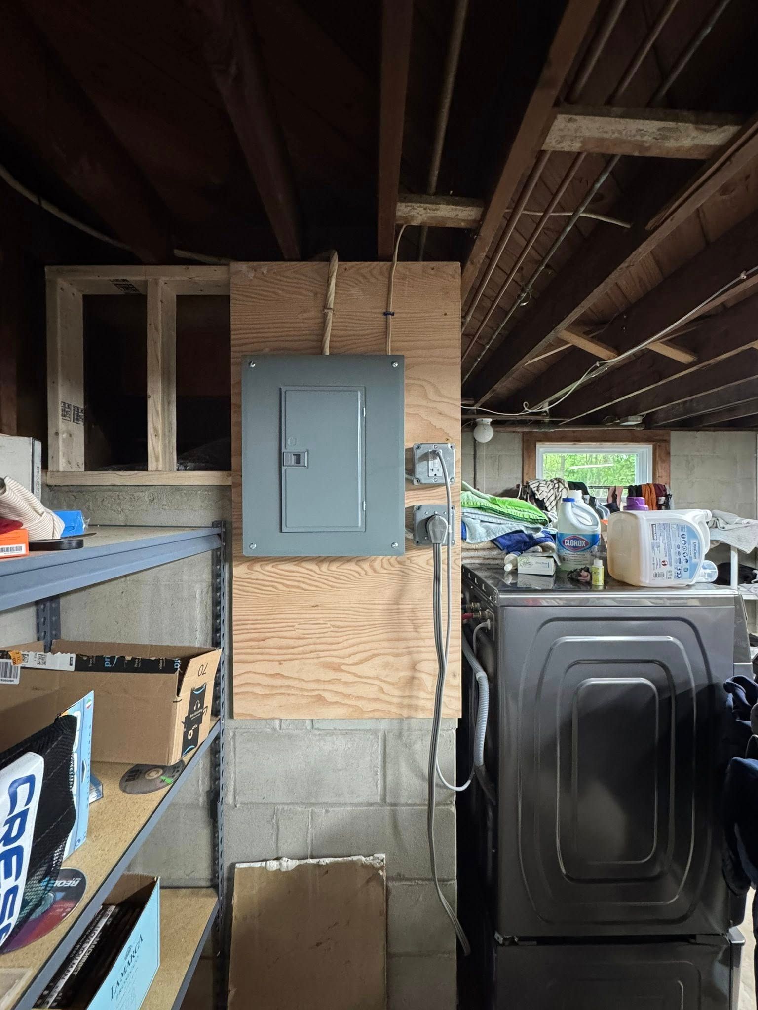 Electrical panel mounted on plywood in a basement; shelves, washing machine, and clutter are also visible.