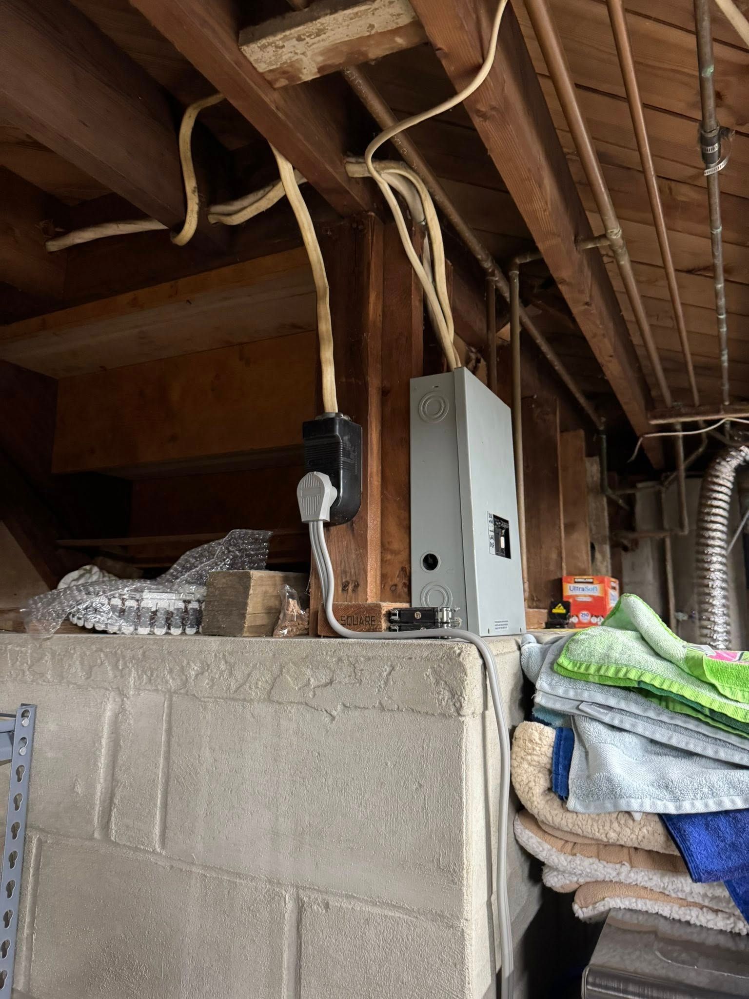 Electrical panel and outlet mounted on a concrete block wall in a basement, with exposed wiring and wooden beams overhead.