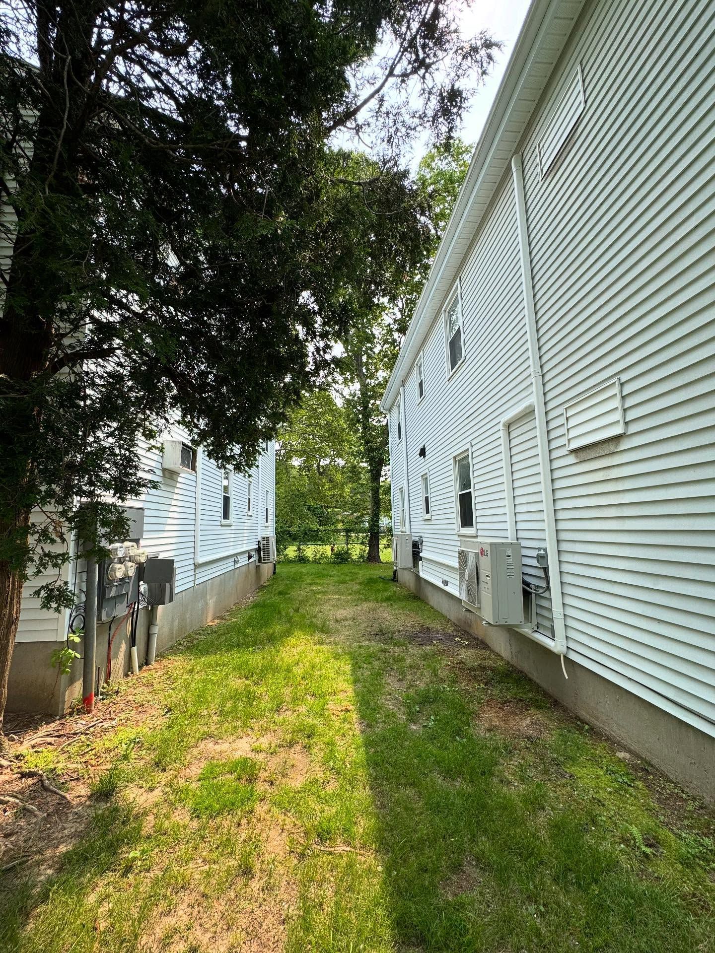 Narrow grassy pathway between two white buildings, with AC units and a tree.