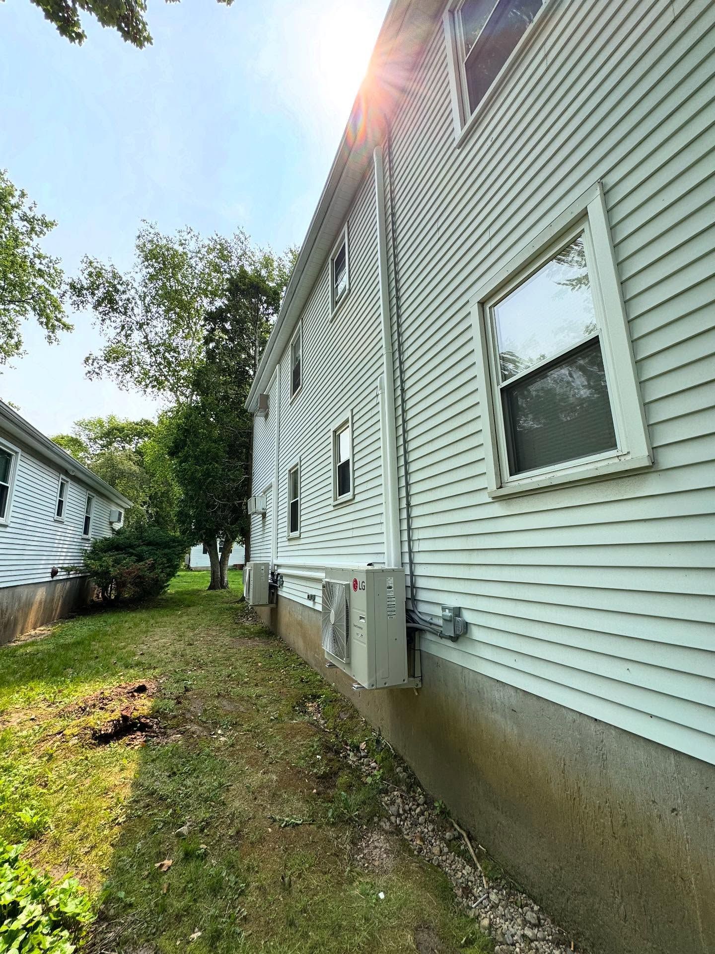 Side of a light-colored house with multiple windows and a grassy yard. An air conditioning unit is mounted on the wall.