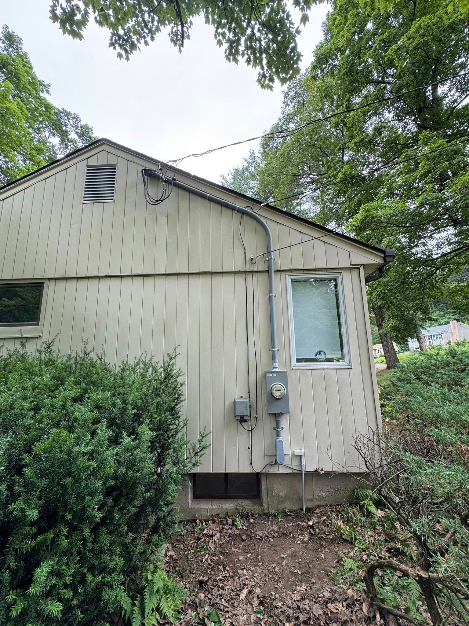 Side of a house with vertical siding, electrical components, a window, and surrounding trees.