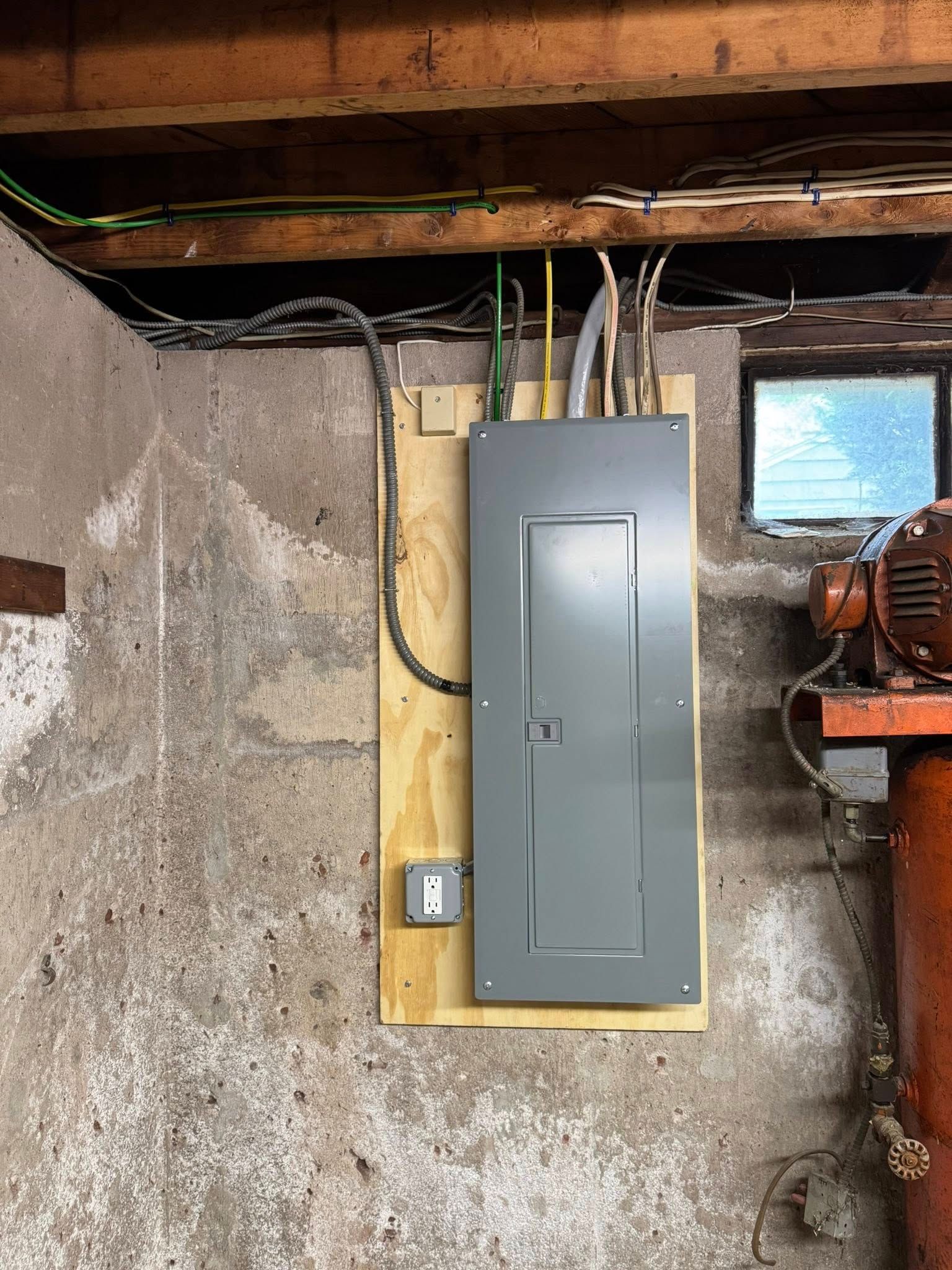Electrical panel mounted on a wooden board on a concrete wall. Cables run above. A small window is to the right.