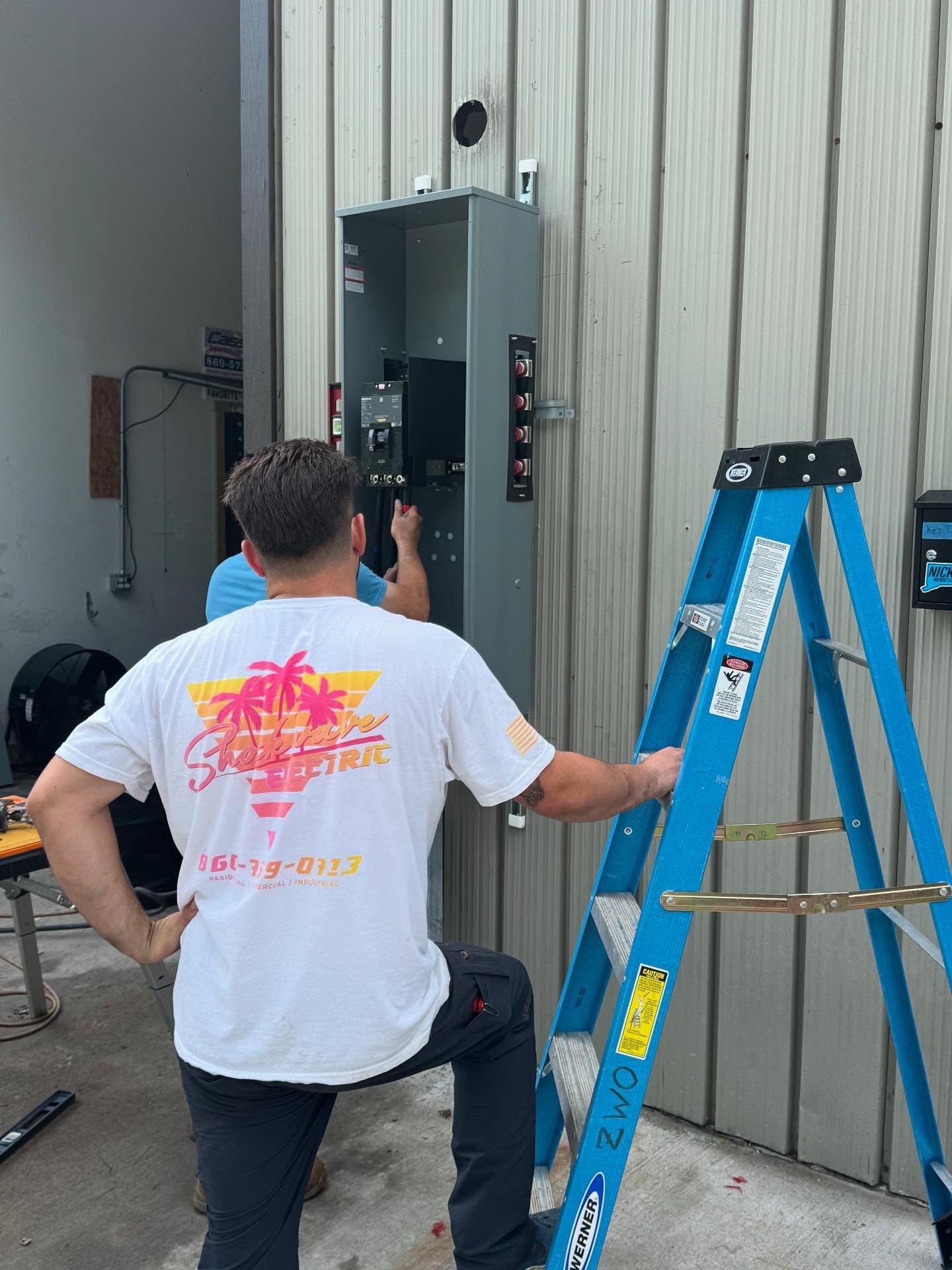 Electricians installing electrical panel on an exterior building wall. A blue ladder is next to the panel.