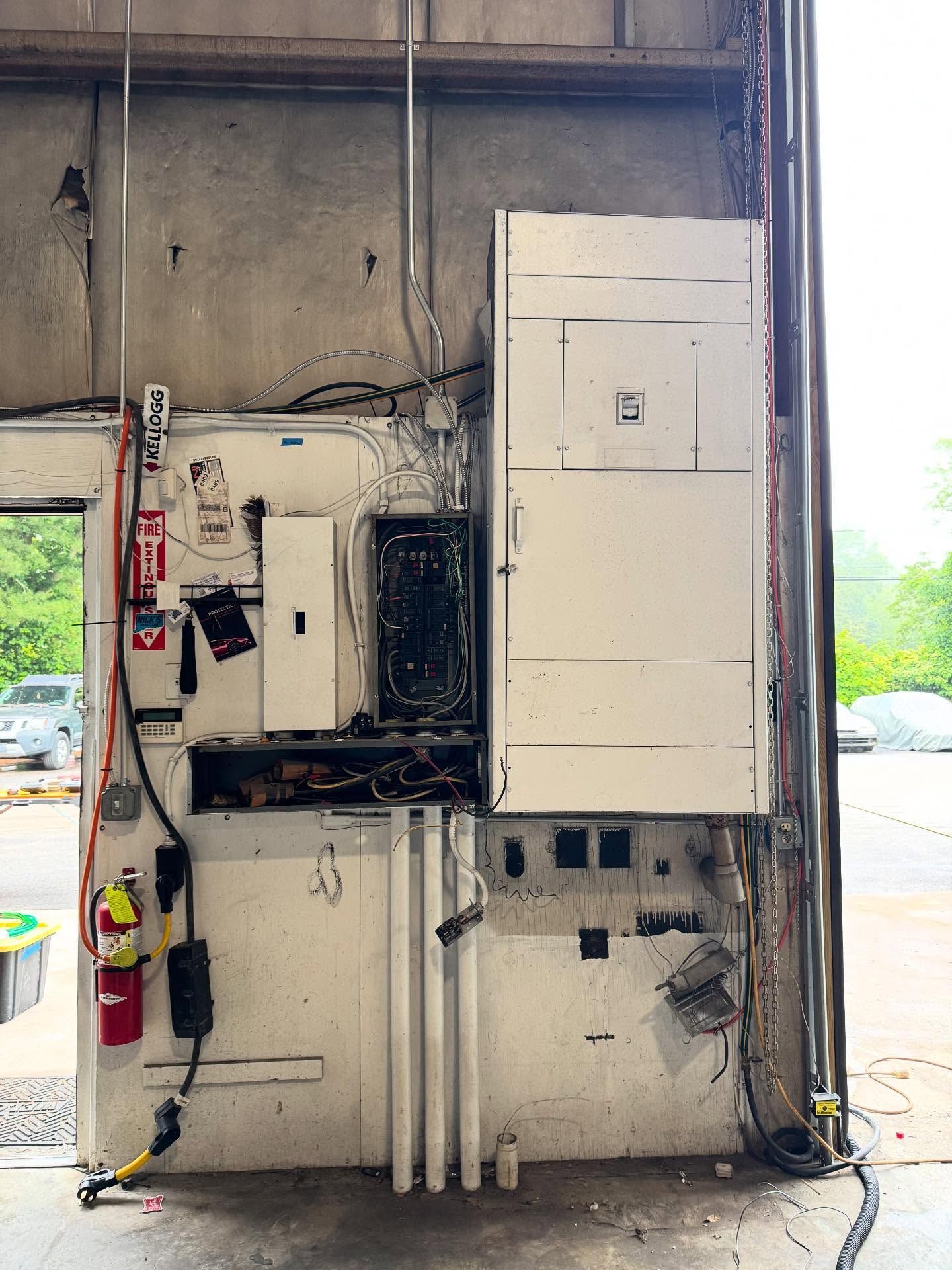 Damaged electrical panel on a concrete wall; fire extinguisher present. Charred panel suggests recent fire.