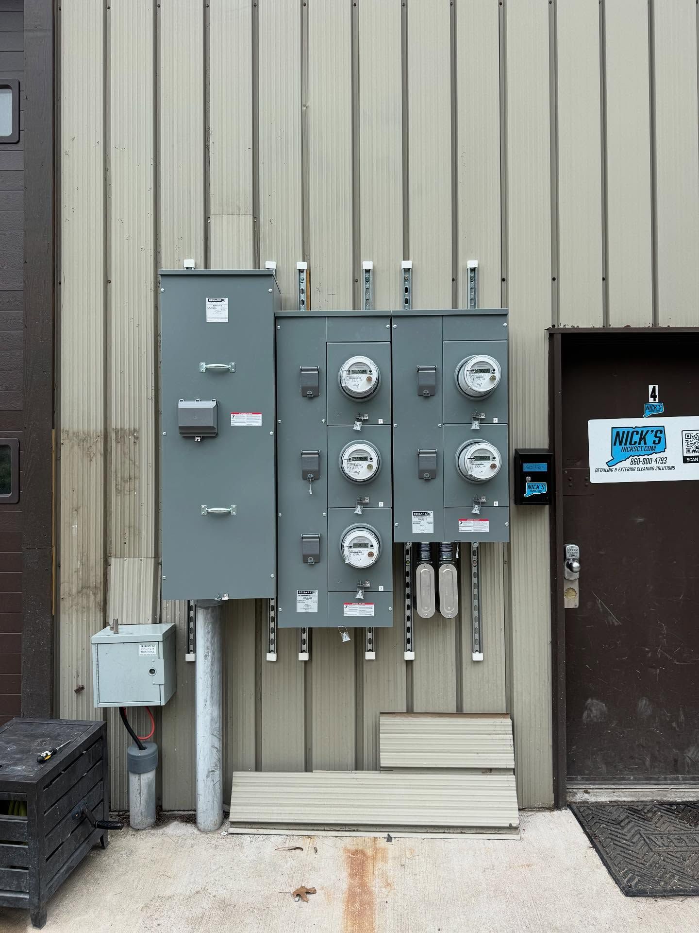 Electrical panel with multiple meters mounted on a corrugated building exterior next to a door.