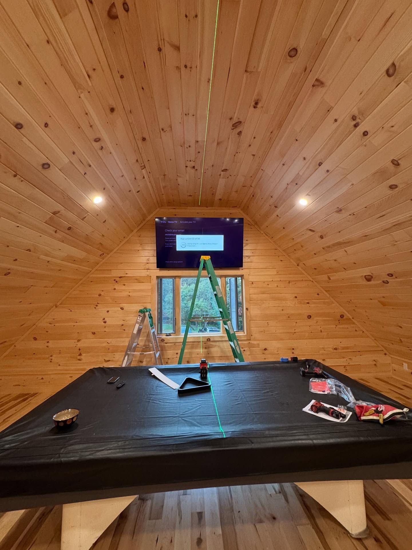 A TV mounted on a wood-paneled wall above a covered pool table. Construction tools and a ladder are present.