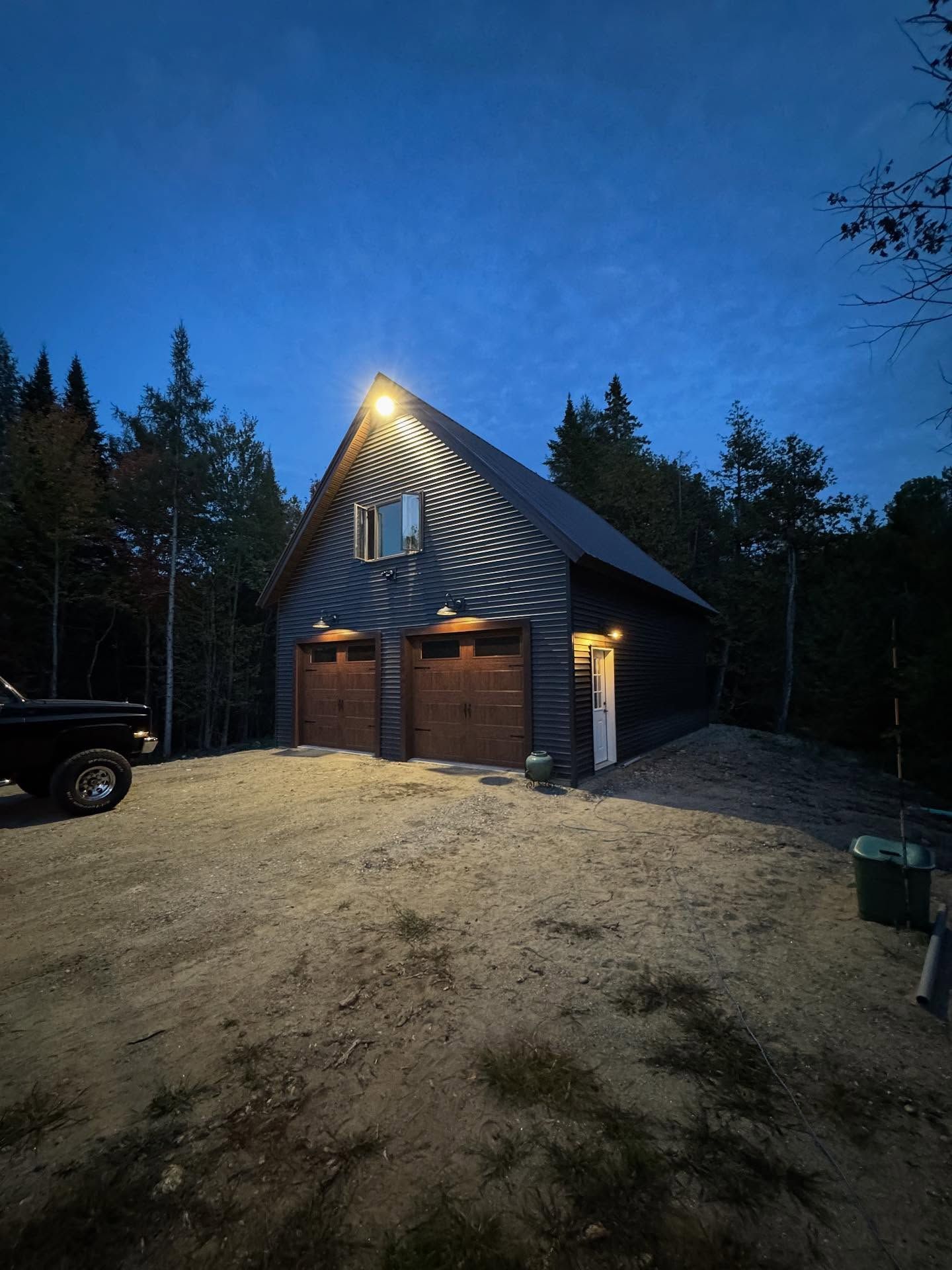Two-bay garage at dusk, lit by overhead floodlight. Wooden doors, small window, gravel driveway.