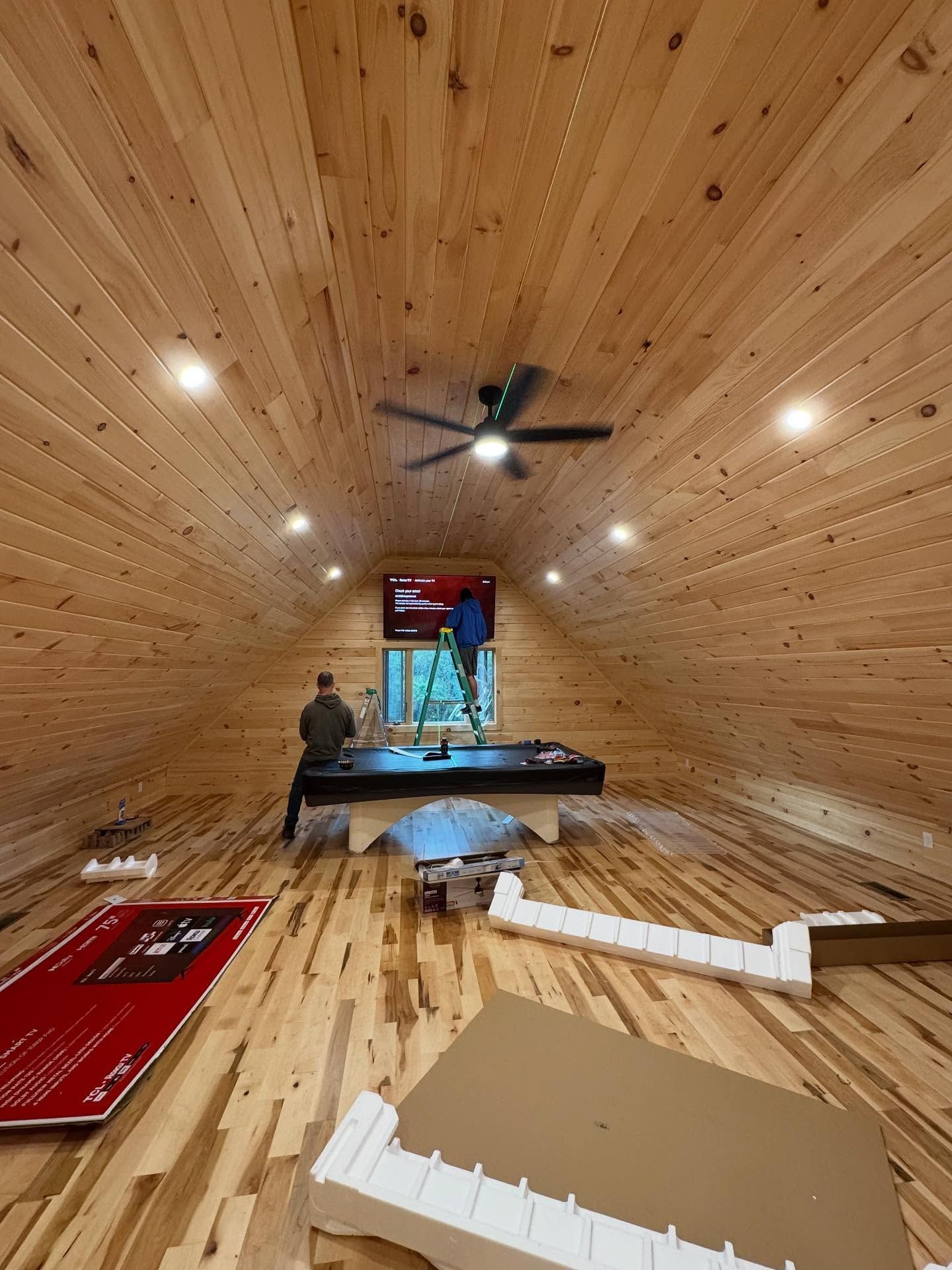 Two people working inside a wood-paneled, vaulted-ceiling room; one on a ladder near a window.