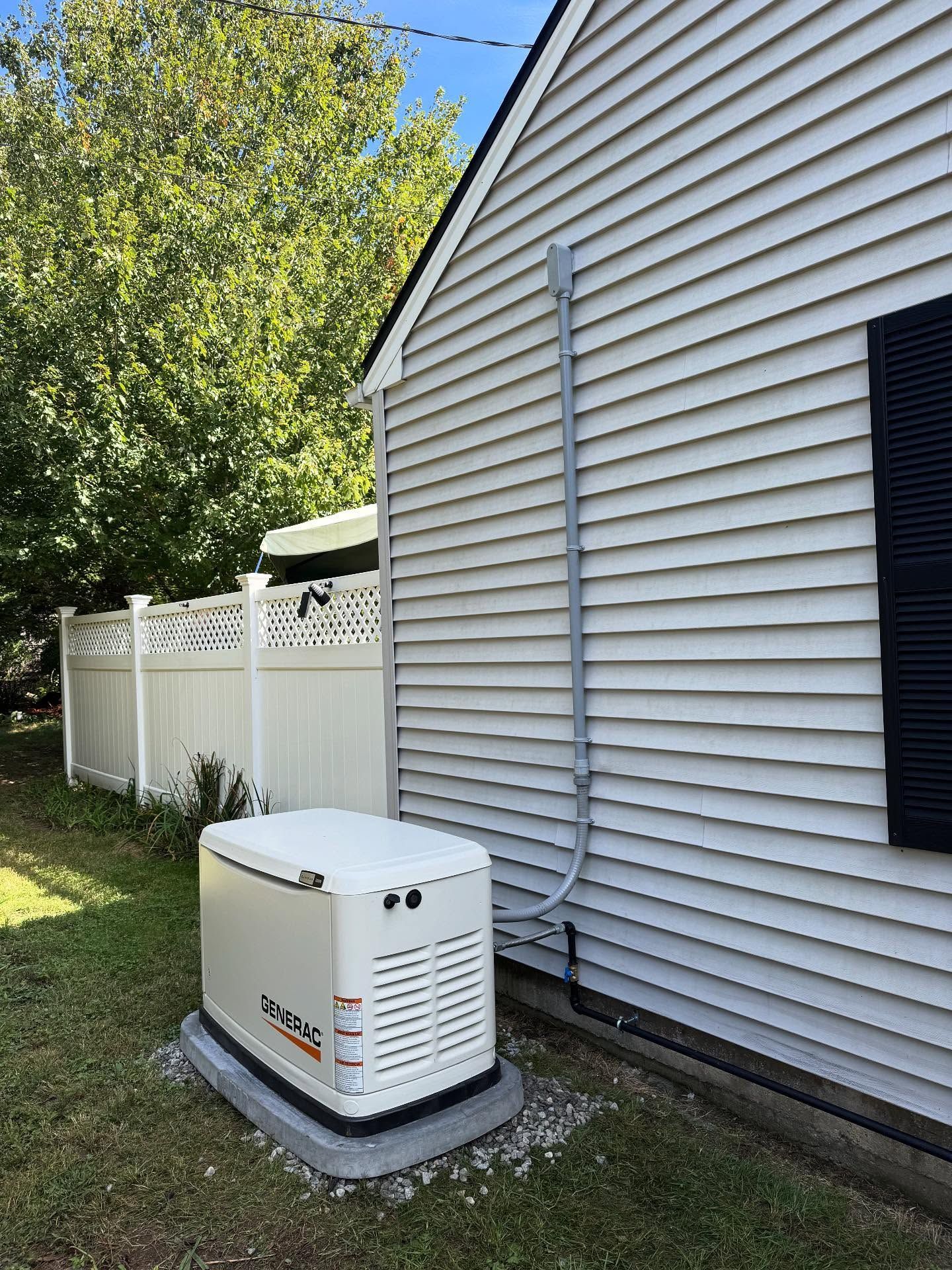 Generator next to a white house with a conduit running up the wall, in front of a white fence.
