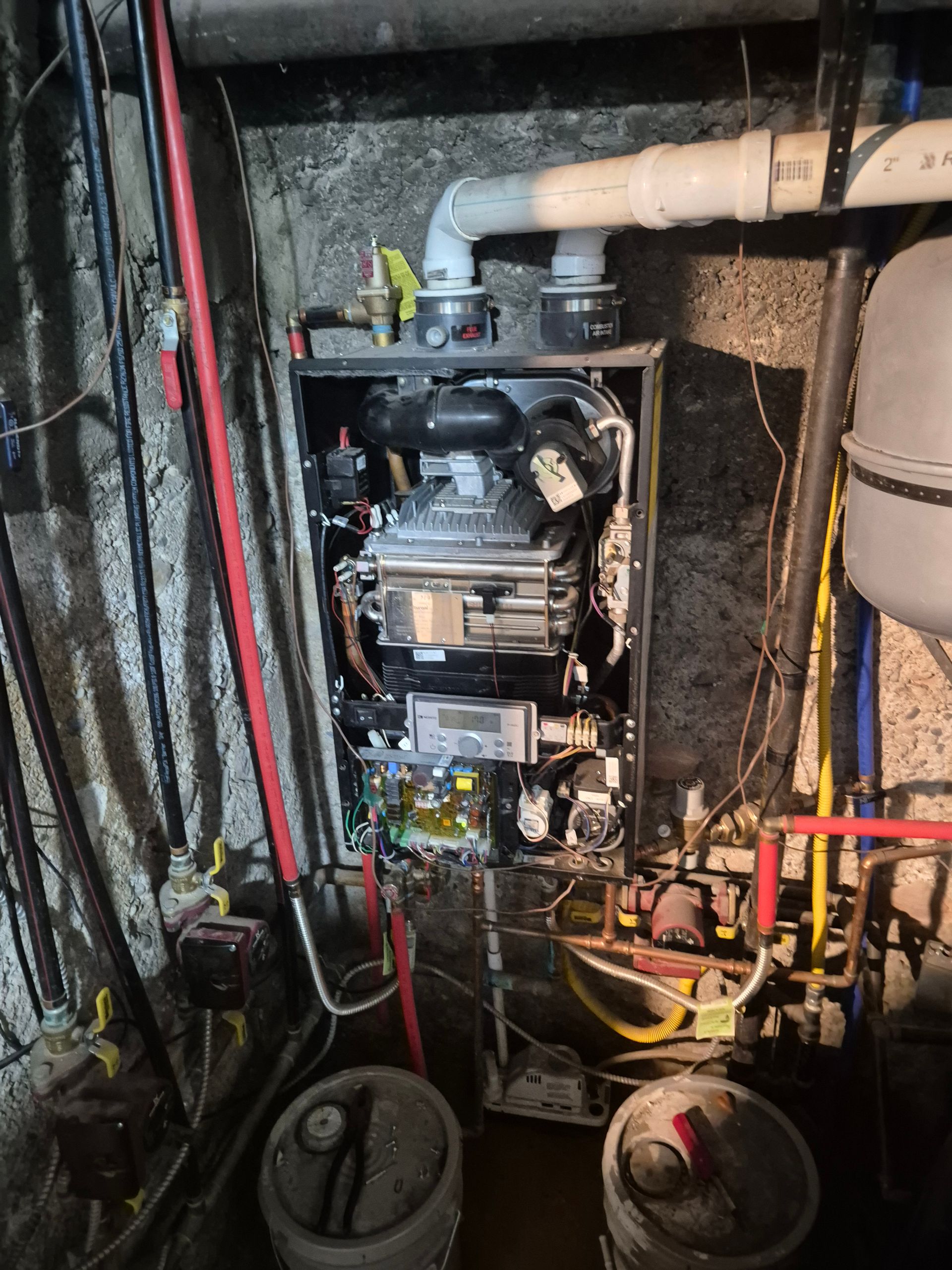 Furnace in a basement; various pipes and wires surround it. Two buckets are in front of the unit.