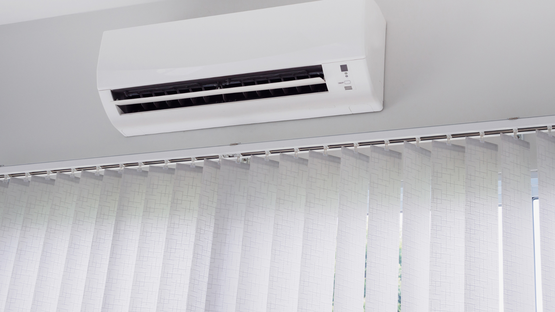 White air conditioner unit mounted on a white ceiling above vertical blinds.