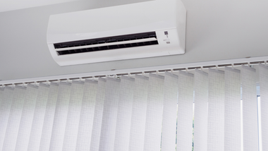 White air conditioner unit mounted on a white ceiling above vertical blinds.