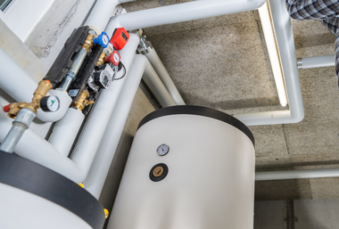Water heater and pipes in a basement. White tank with gauges, surrounded by insulated plumbing.