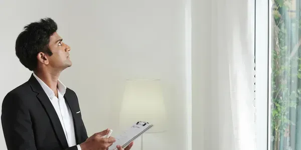 Man in a suit looking up, holding a clipboard, standing near a window.