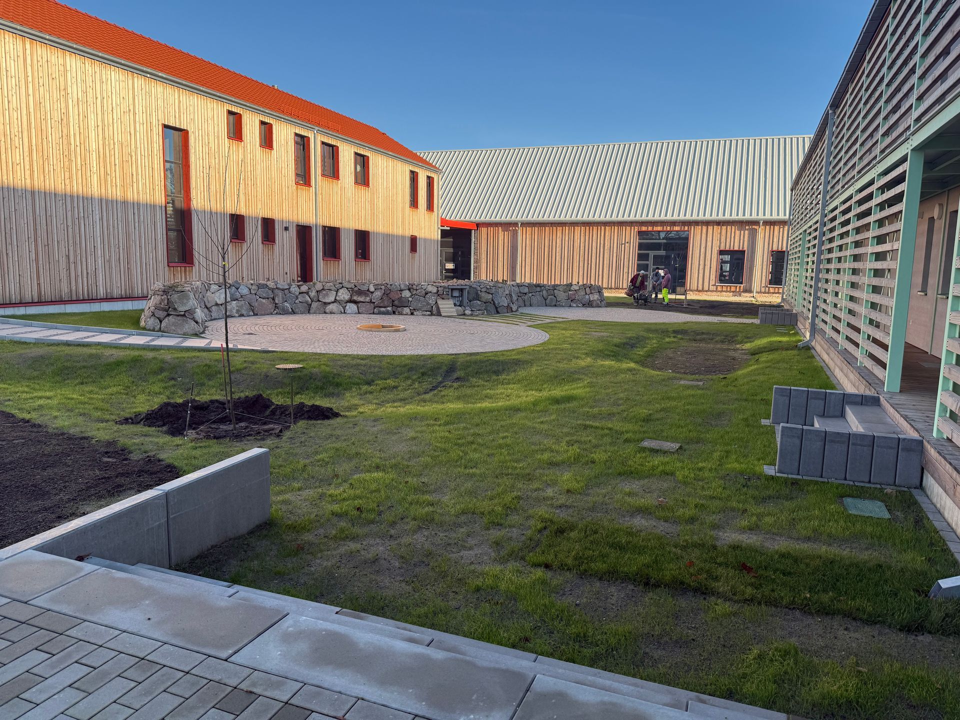 Innenhof mit modernen Holzbauten, grünem Rasen und einer steinernen Sitzgruppe. Klarer Himmel.