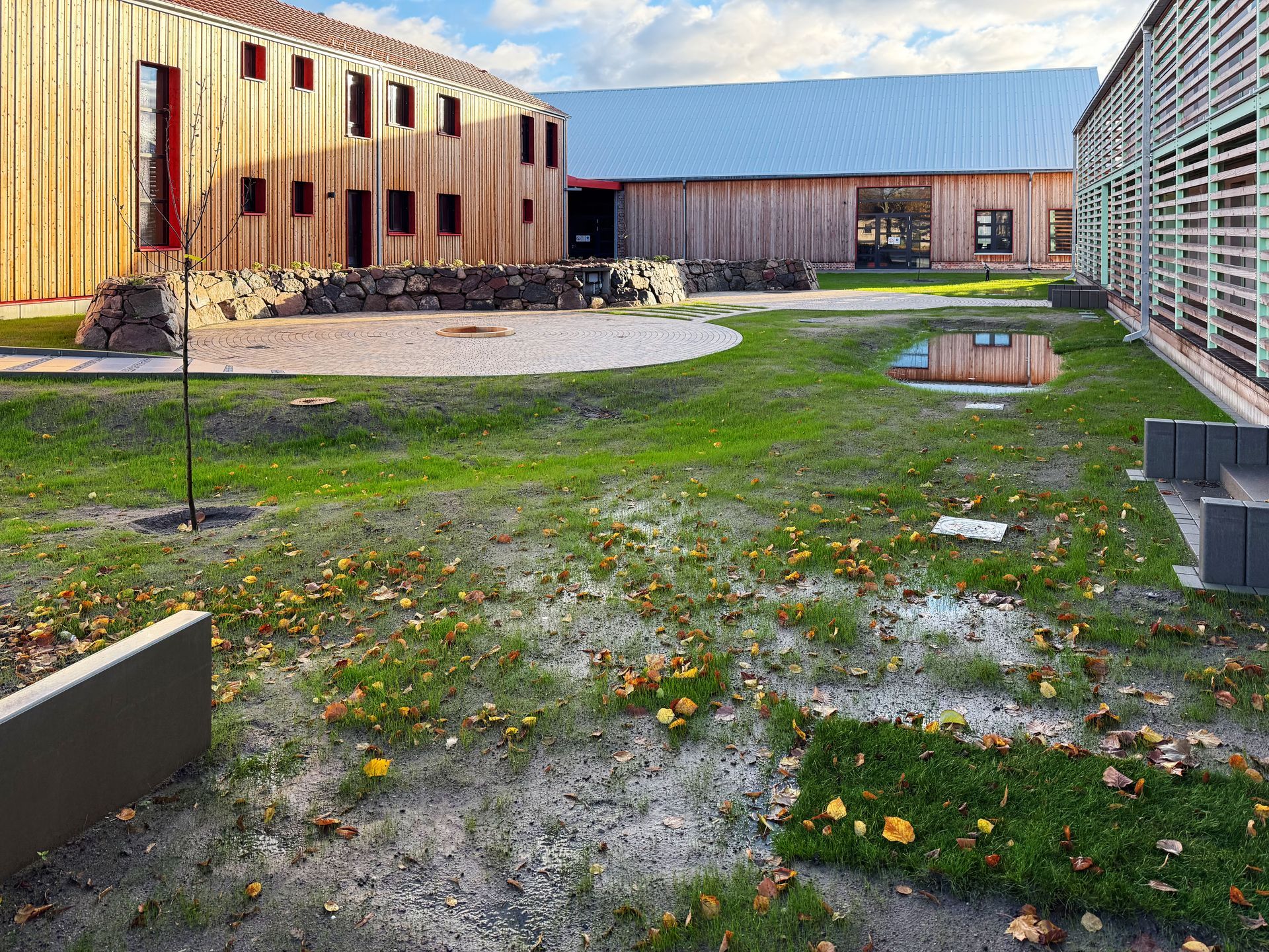 Holzbauten umgeben einen grasbewachsenen, teilweise überfluteten Hof mit herabgefallenen Blättern. Strahlend heller Himmel.