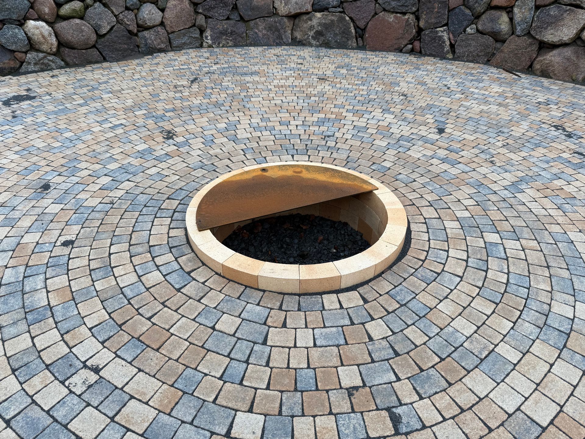 Runde, gemauerte Feuerstelle mit offenem Deckel, umgeben von Kopfsteinpflaster.