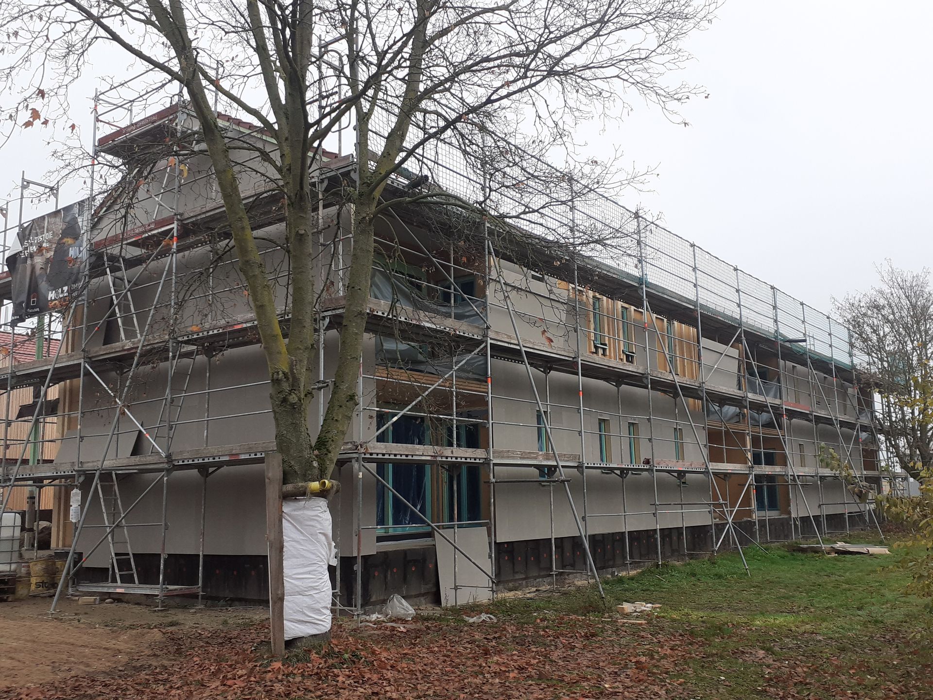 Ein großes im Bau befindliches Gebäude mit Gerüst darum und einem Baum davor.