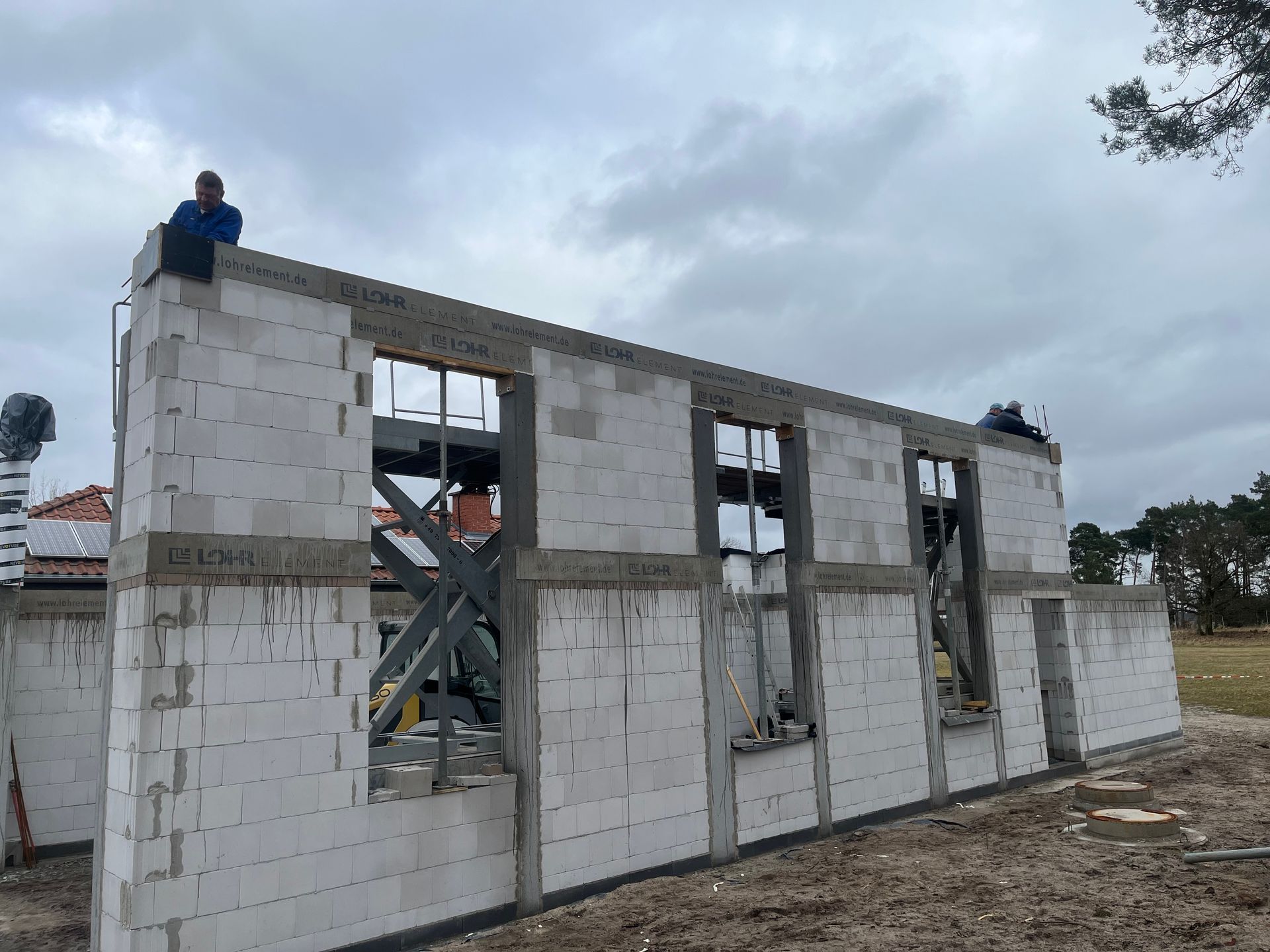 Baustelle: Teilweise errichtetes Haus mit Betonsteinwänden, Fensterrahmen und Arbeitern auf dem Dach.